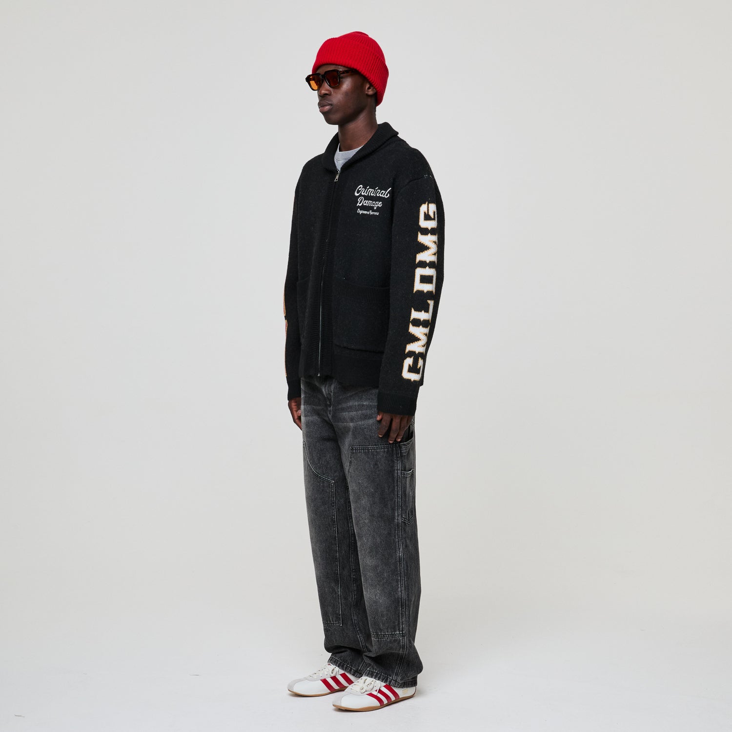 Person wearing a Criminal Damage black hoodie with text, gray jeans, and red cap on a white background