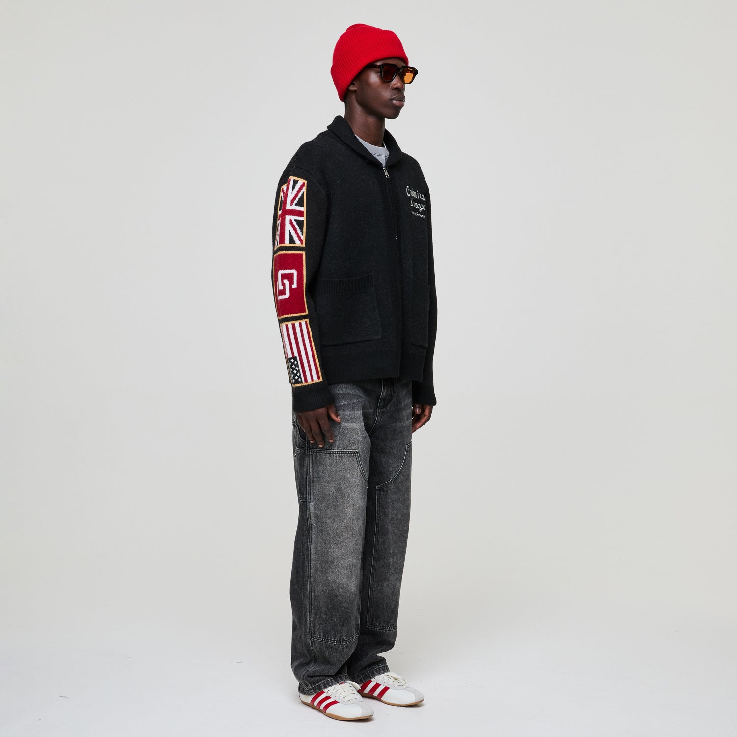 Person wearing a Criminal Damage black hoodie with a Union Jack design, gray jeans, and a red cap on a white background