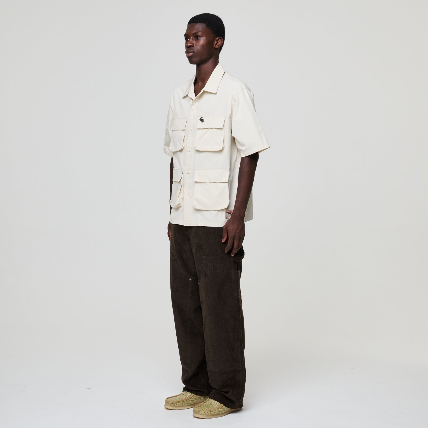 Person wearing a beige shirt with multiple pockets and dark pants on a white background