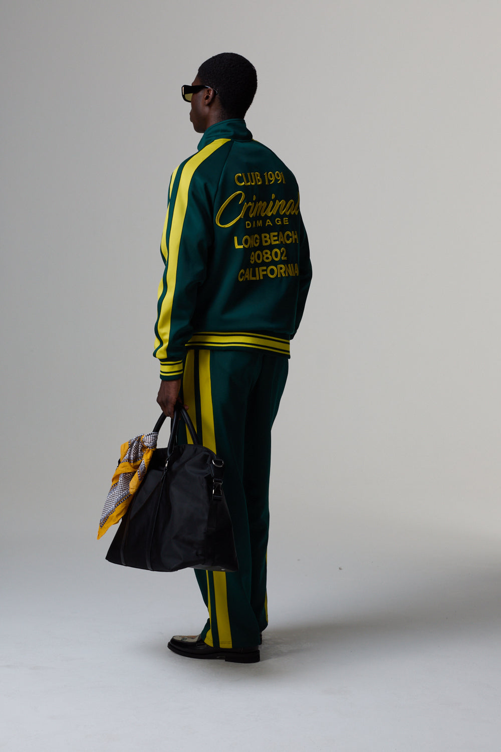 Model wearing a Criminal Damage green and yellow tracksuit with text, holding a bag on a plain background