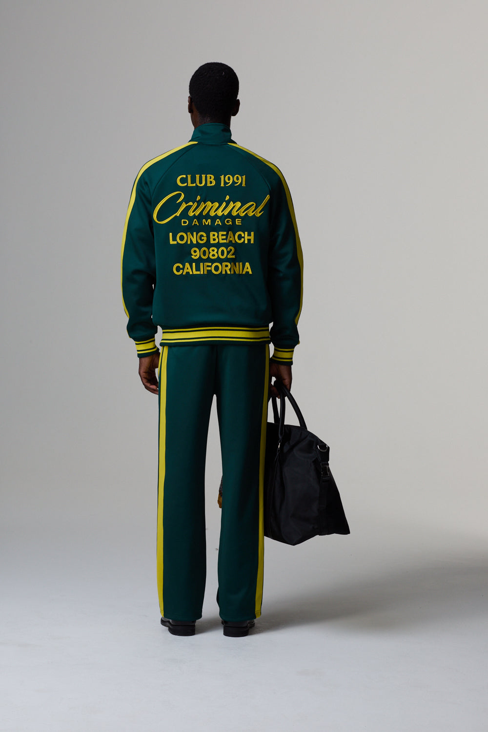 Model wearing a Criminal Damage green and yellow tracksuit with text on the back, holding a black bag.