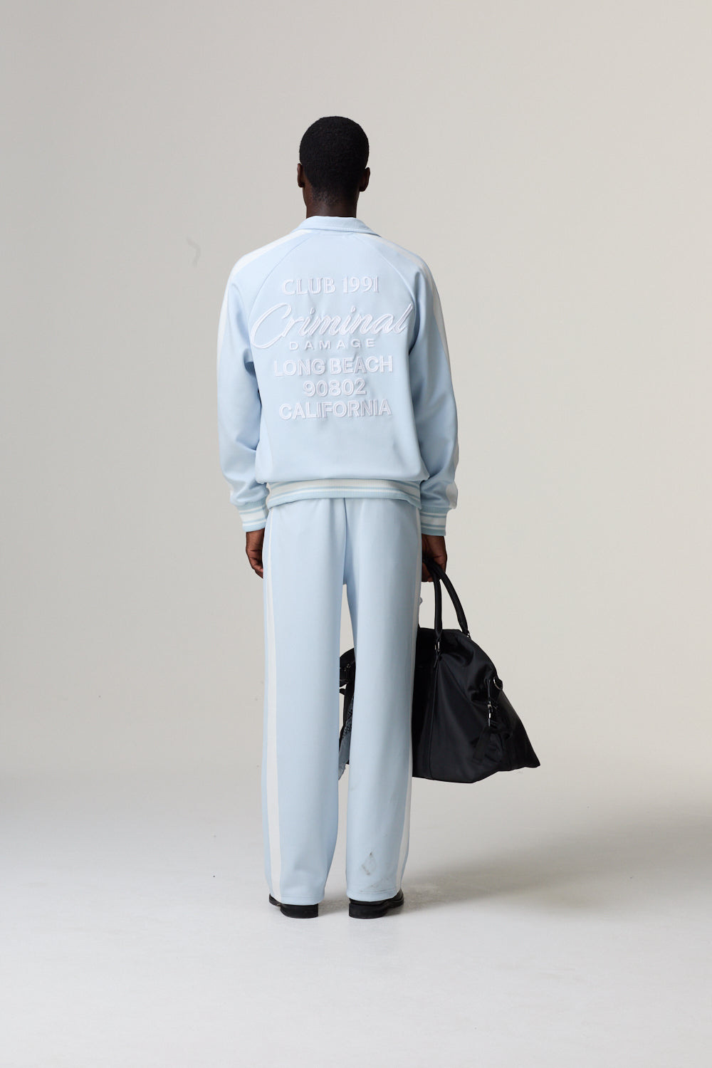 Model wearing a Criminal Damage light blue tracksuit holding a black bag 
