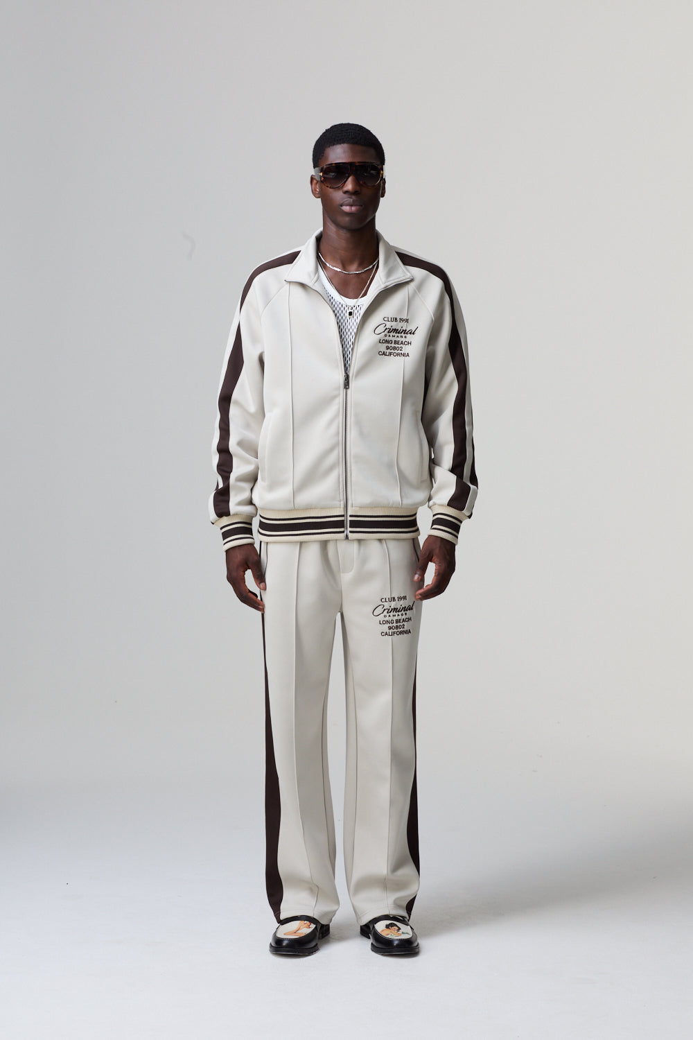 Model wearing a Criminal Damage white tracksuit with brown stripes on a grey background