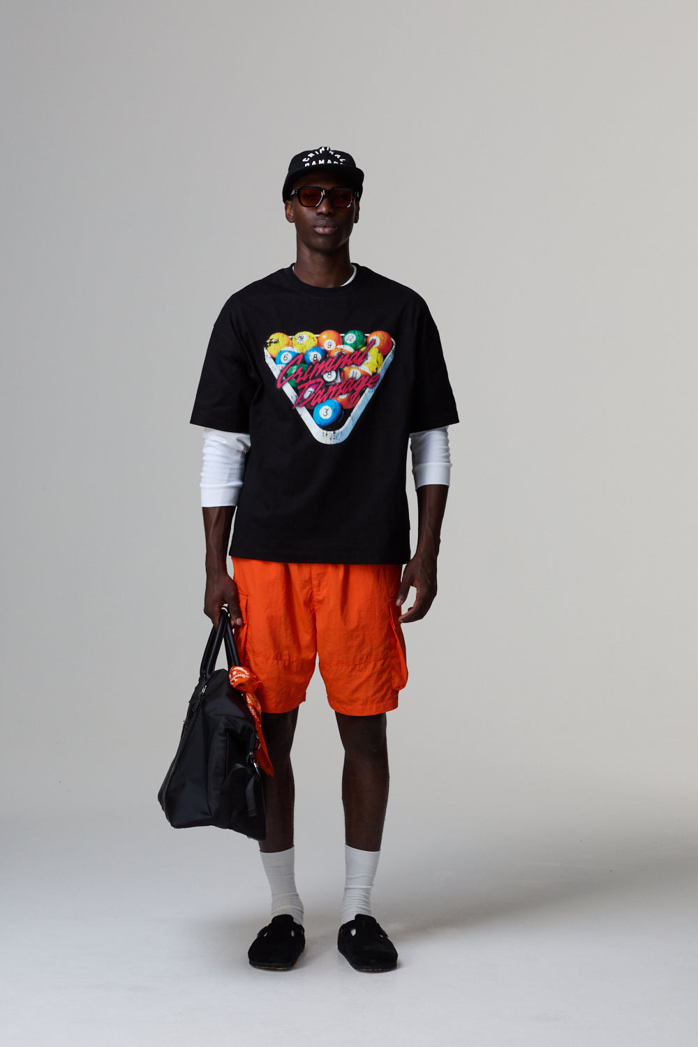 Model wearing a black Criminal Damage t-shirt with graphic design, orange shorts, and holding a black bag on a grey background