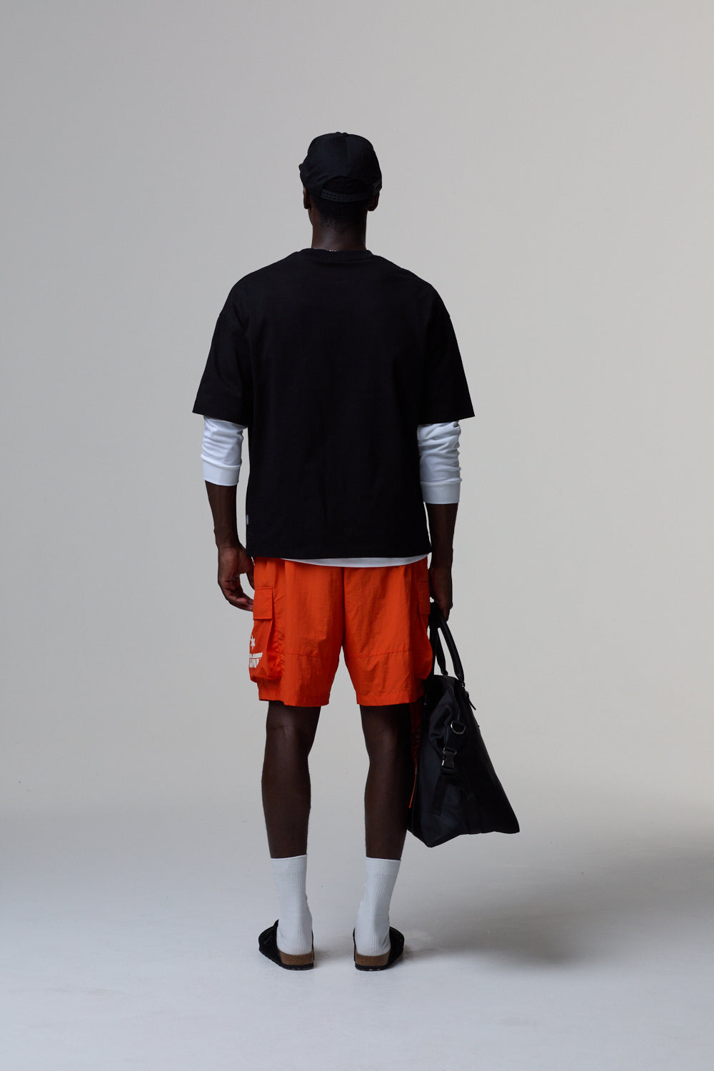 Model wearing a black Criminal Damage t-shirt, orange shorts, and white socks, holding a black bag on a grey background