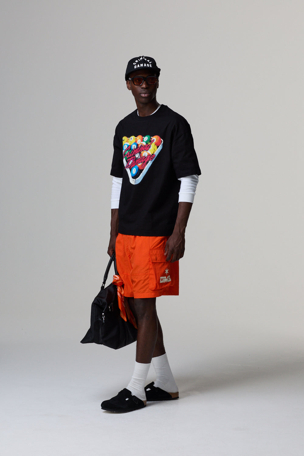 Model wearing a black Criminal Damage t-shirt with colourful design, orange shorts, and black shoes on a grey background