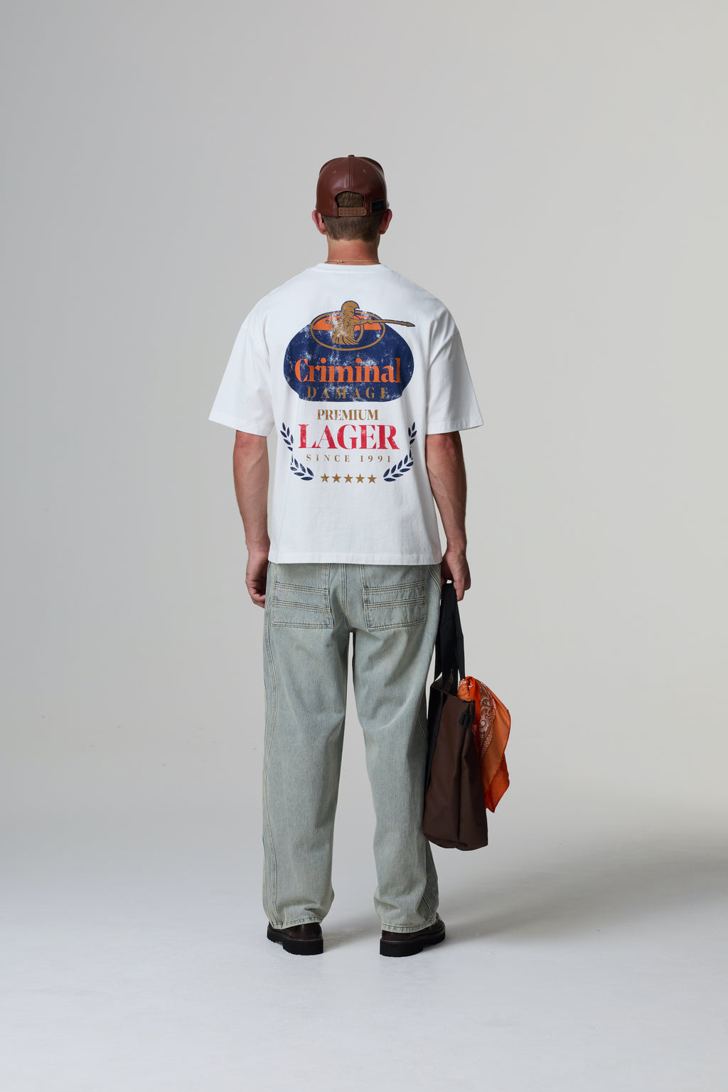 Model wearing a white Criminal Damage t-shirt with a graphic design, standing against a grey background