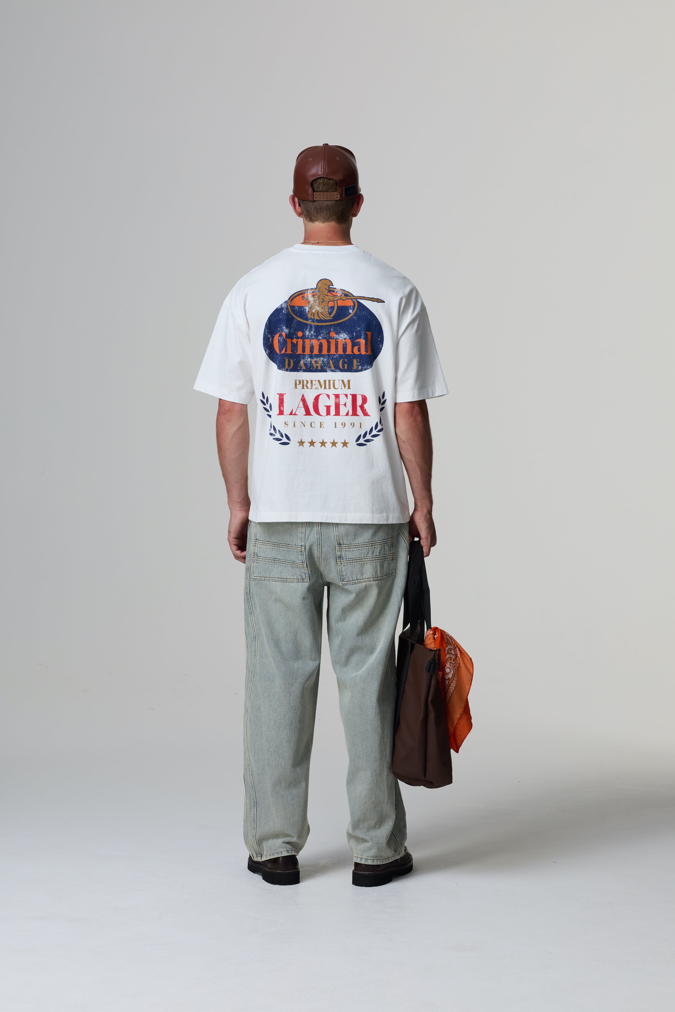 Model wearing a white Criminal Damage t-shirt with a graphic design, standing against a grey background