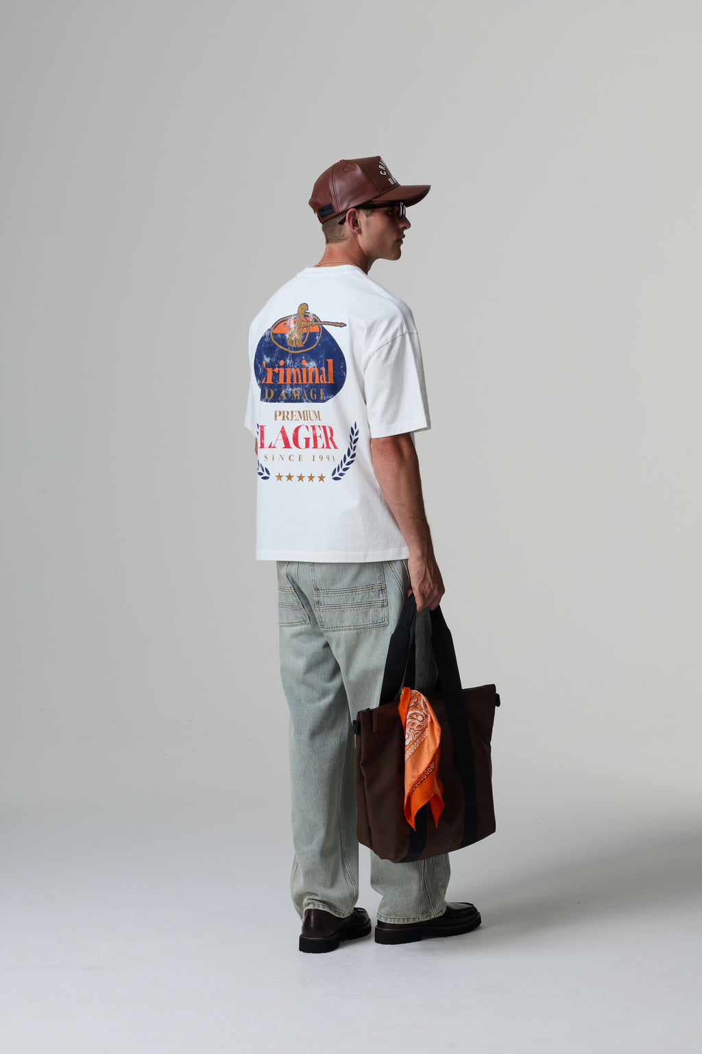 Model wearing a white Criminal Damage t-shirt with a graphic design, holding a bag, on a grey background.