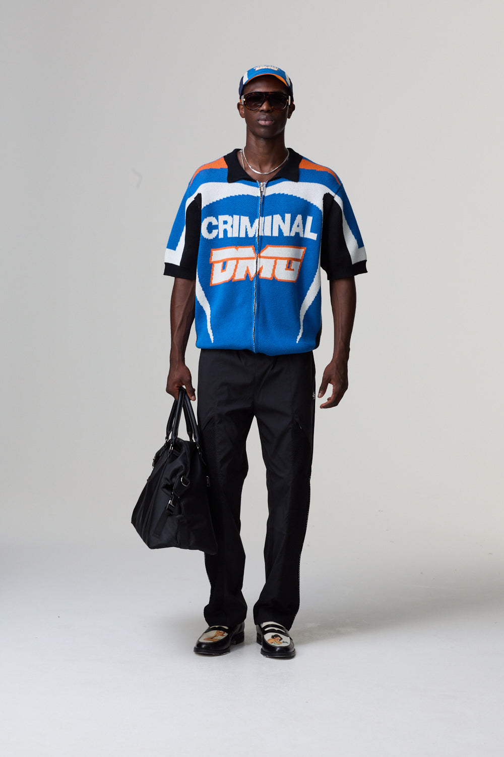 Model wearing a blue and white jacket with 'Criminal Damage' text, holding a black bag on a grey background