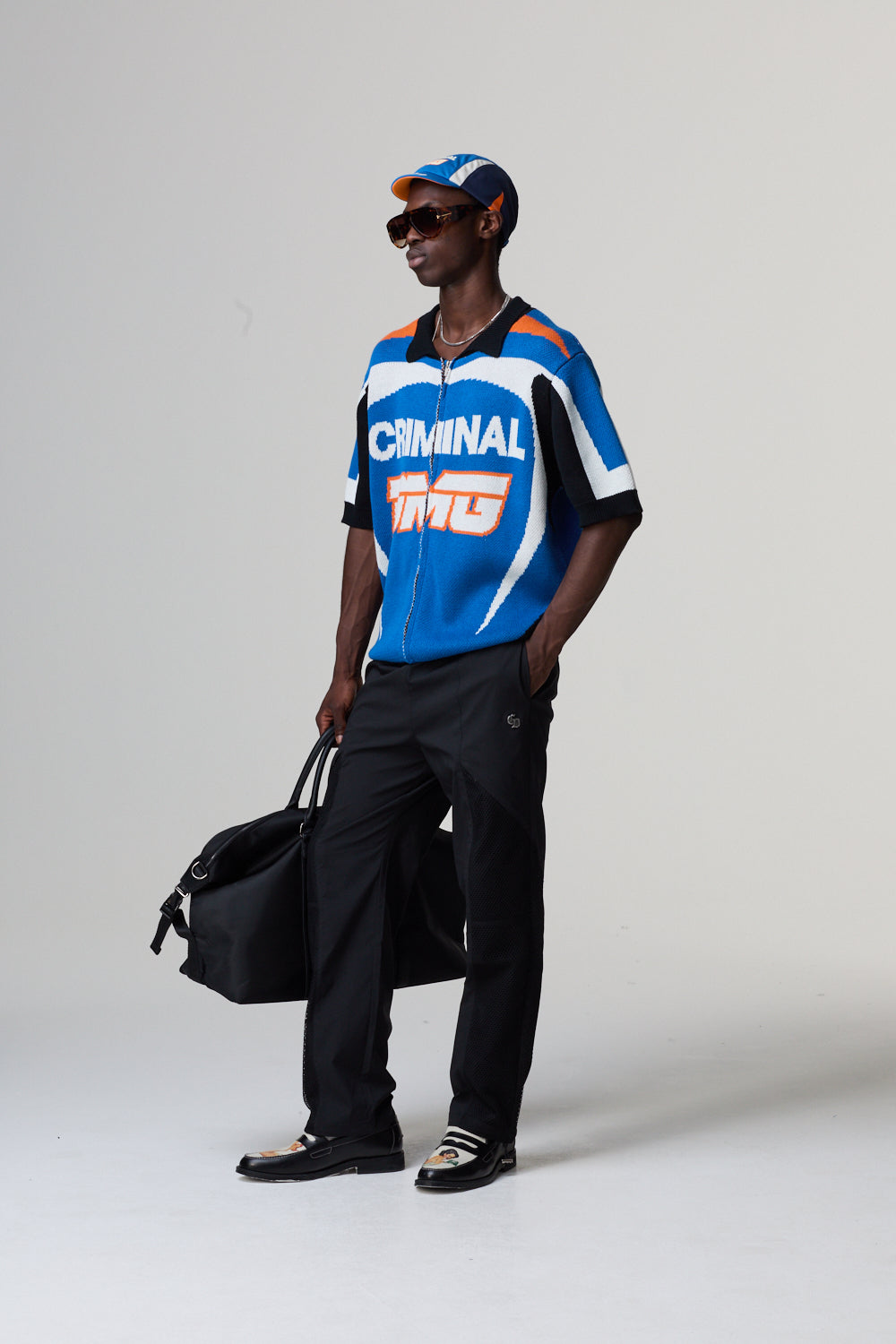 Model wearing a Criminal Damage blue and orange sports jersey with 'Criminal Damage' branding, holding a black bag on a grey background.