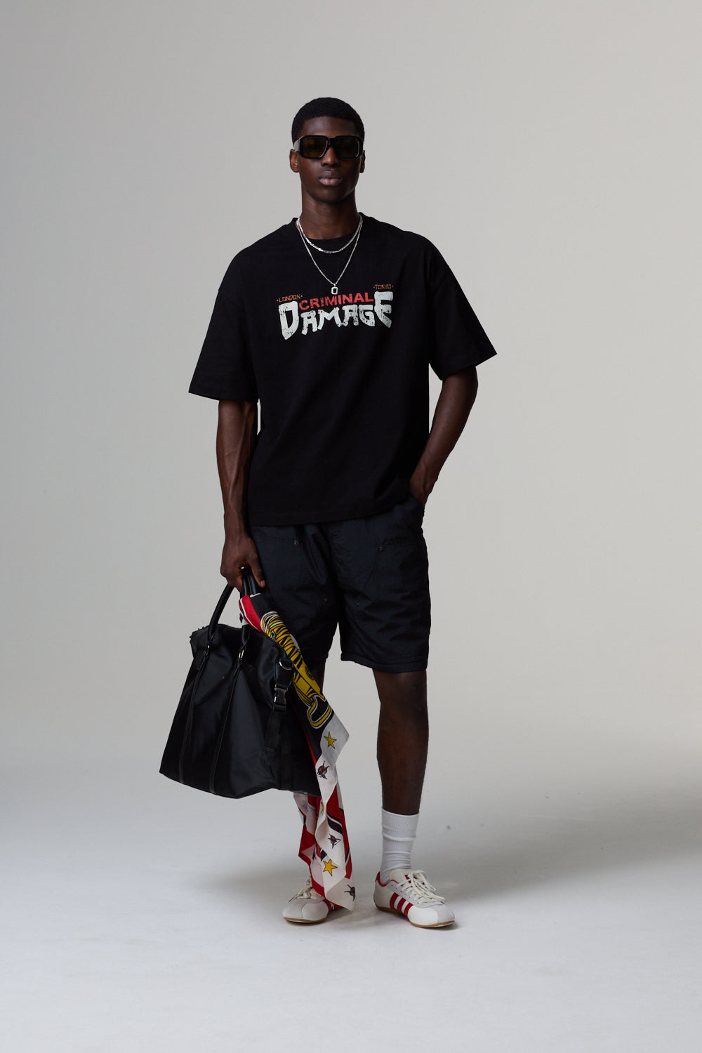 Model wearing a black Criminal Damage t-shirt with text, holding a bag and colourful shoes on a grey background