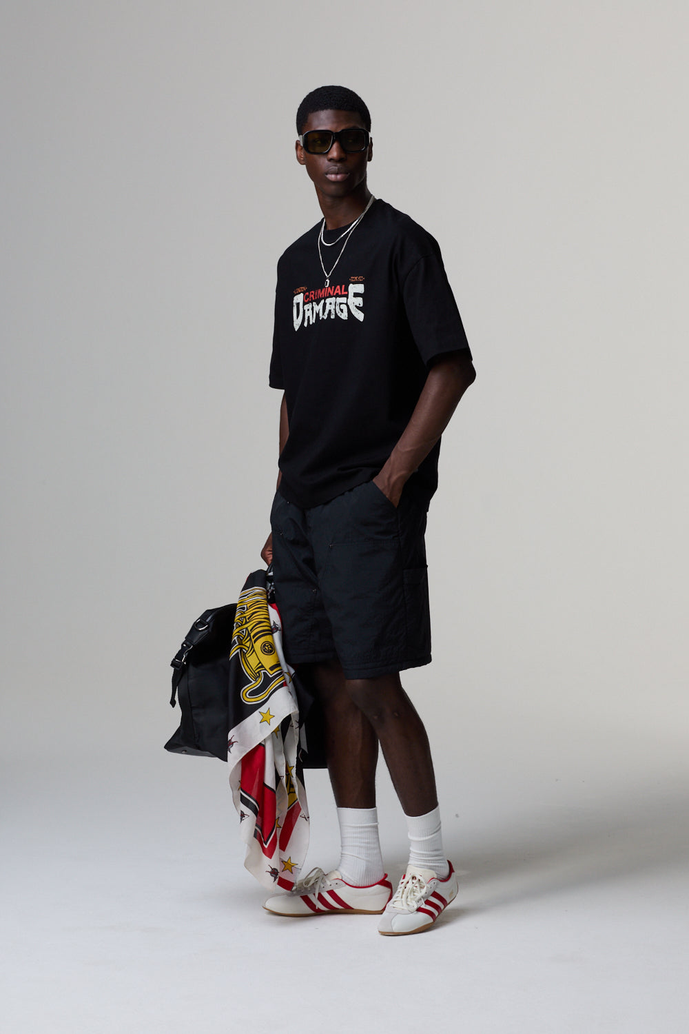 Model wearing a black Criminal Damage t-shirt with a logo, black shorts, white socks, and red sneakers, holding a backpack.