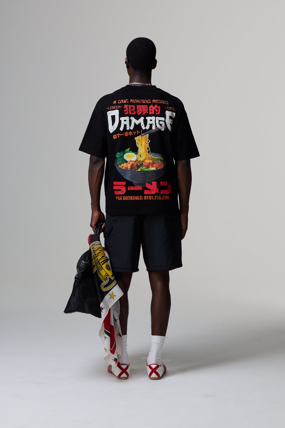 Model wearing a Criminal Damage black t-shirt with colourful graphics and text, holding a jacket on a grey background