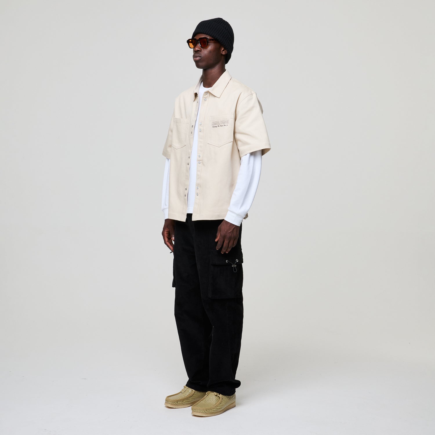 Man wearing a Criminal Damage  beige shirt, black pants, and a black beanie on a white background