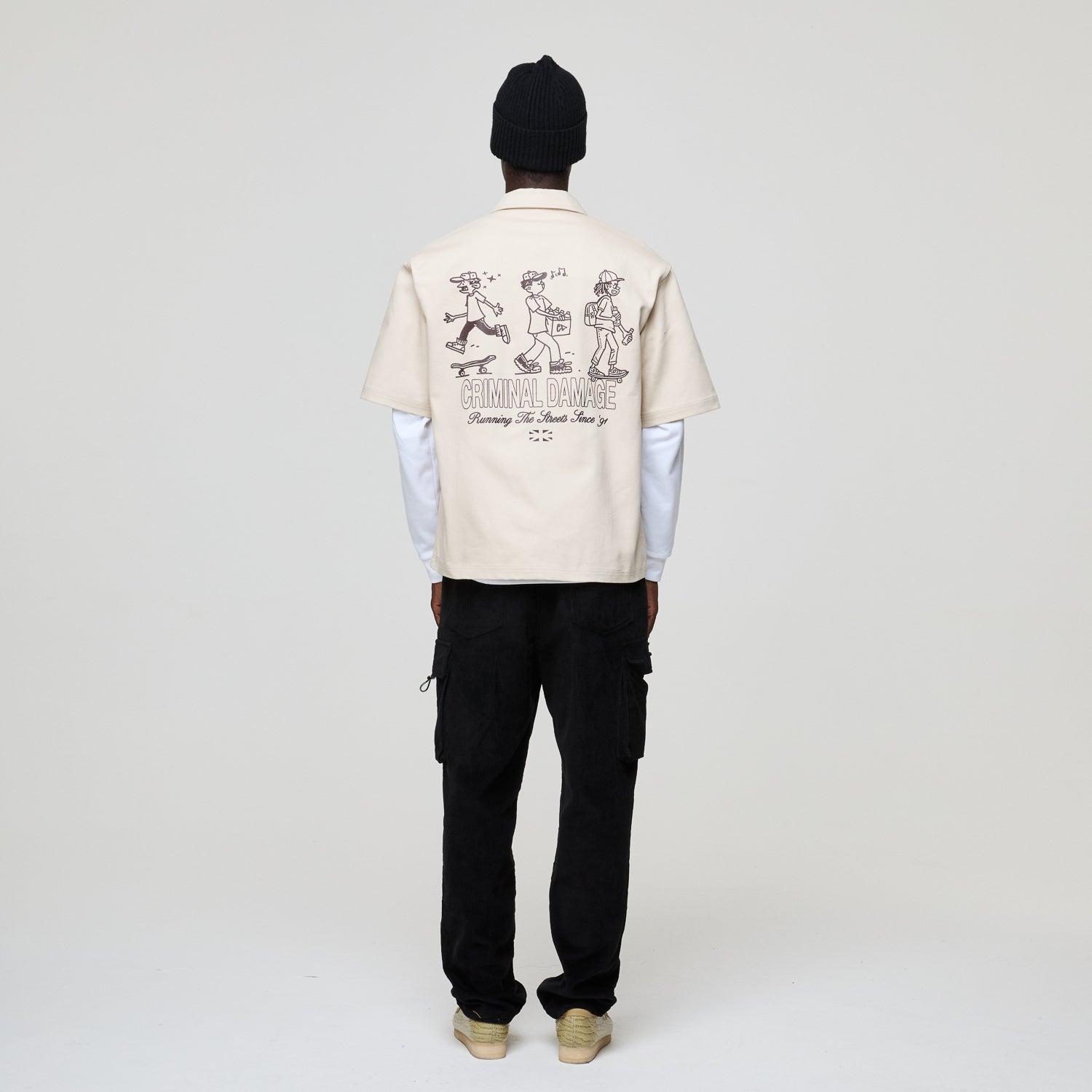 Person wearing a beige Criminal Damage shirt with a graphic design on the back against a white background
