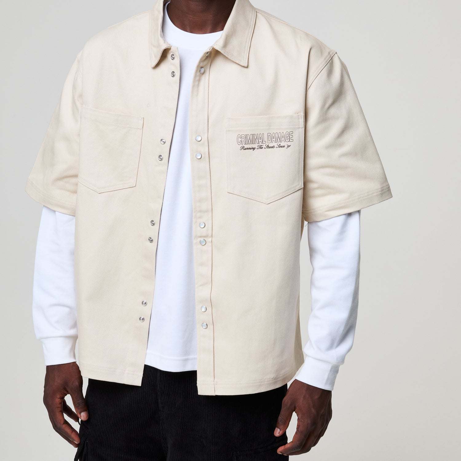 Person wearing a beige Criminal Damage short-sleeve shirt over a white long-sleeve shirt on a plain background