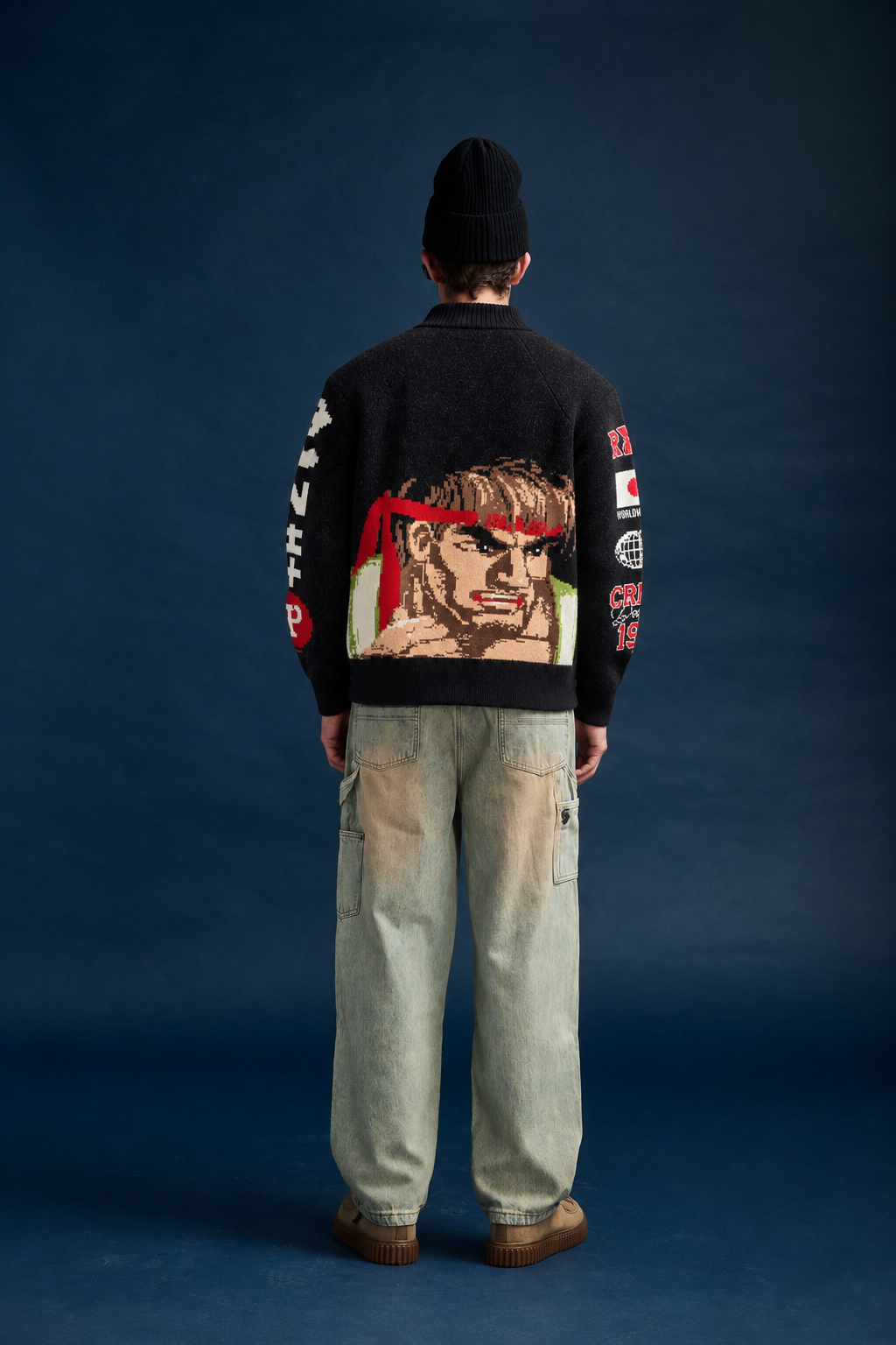 A model shown from the back wearing a black Criminal Damage x Street Fighter knit cardigan with a large graphic of a face and red headband on the back, paired with light-wash jeans, a black beanie, and brown shoes, set against a dark blue studio background.