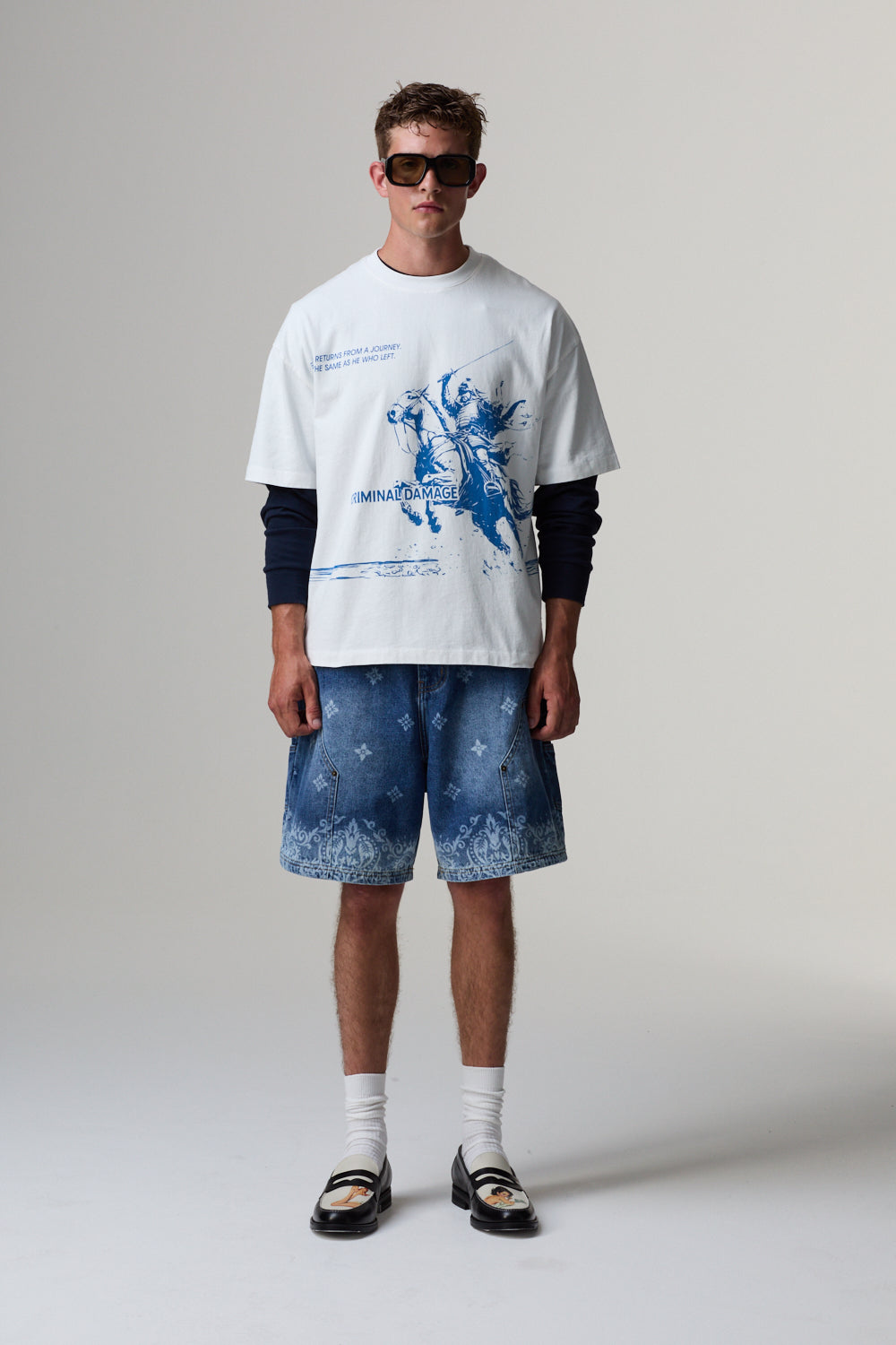 Model wearing a white Criminal Damage t-shirt with a graphic design and blue shorts on a grey background