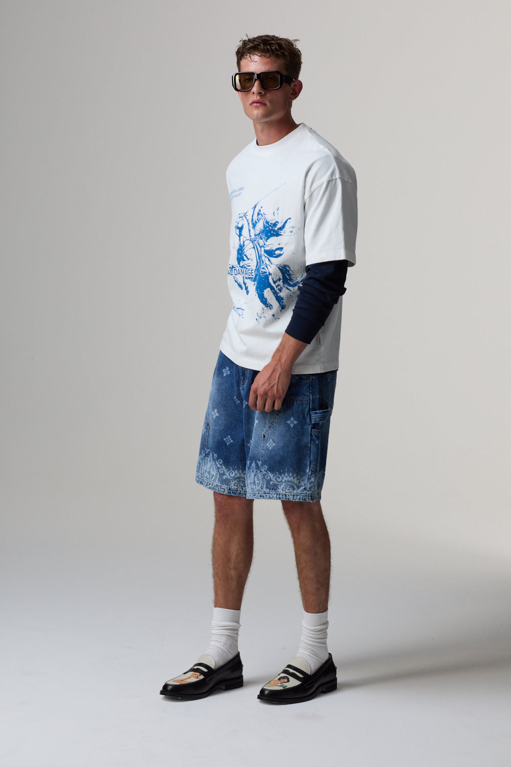Model wearing a white Criminal Damage t-shirt with blue graphic, denim shorts, and black shoes on a grey background