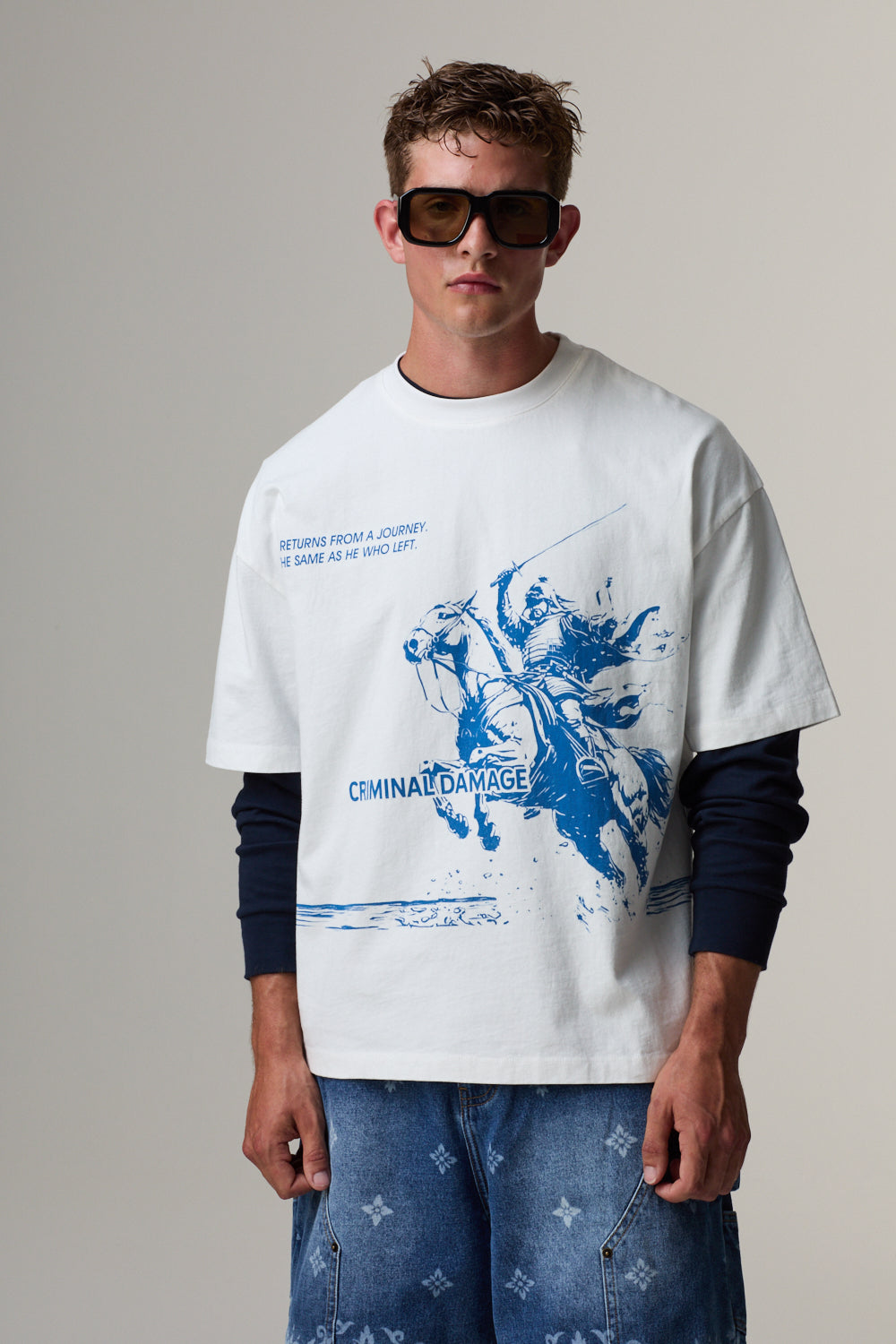 Model wearing a white Criminal Damage t-shirt with blue graphic design and sunglasses on a grey background