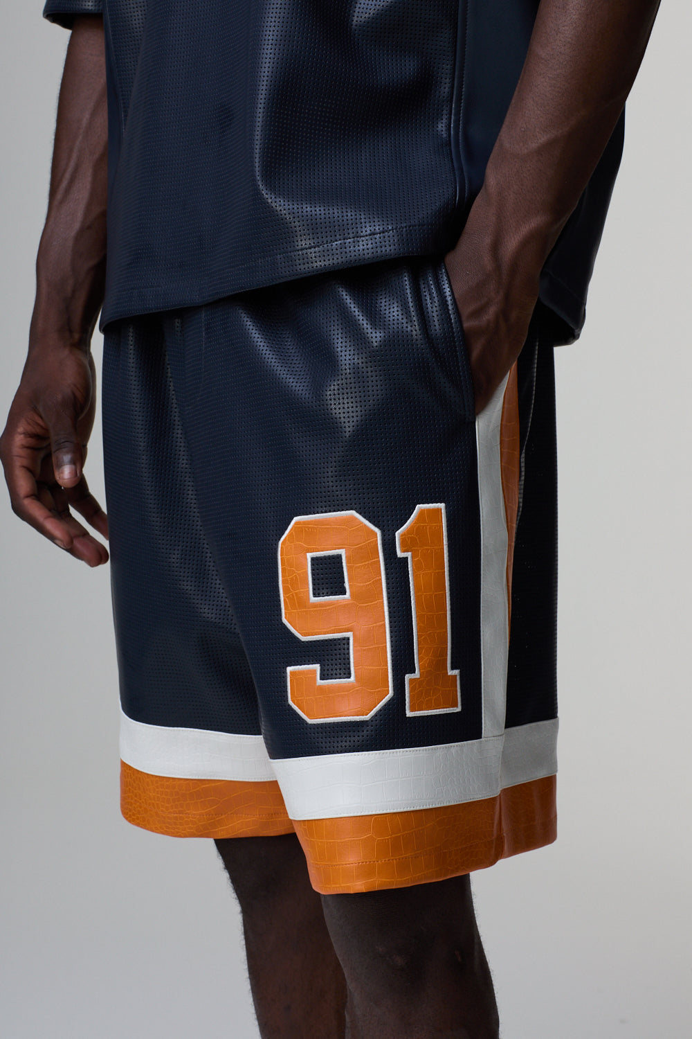Model wearing Criminal Damage navy athletic shorts with orange and white accents and the number 91 on a gray background