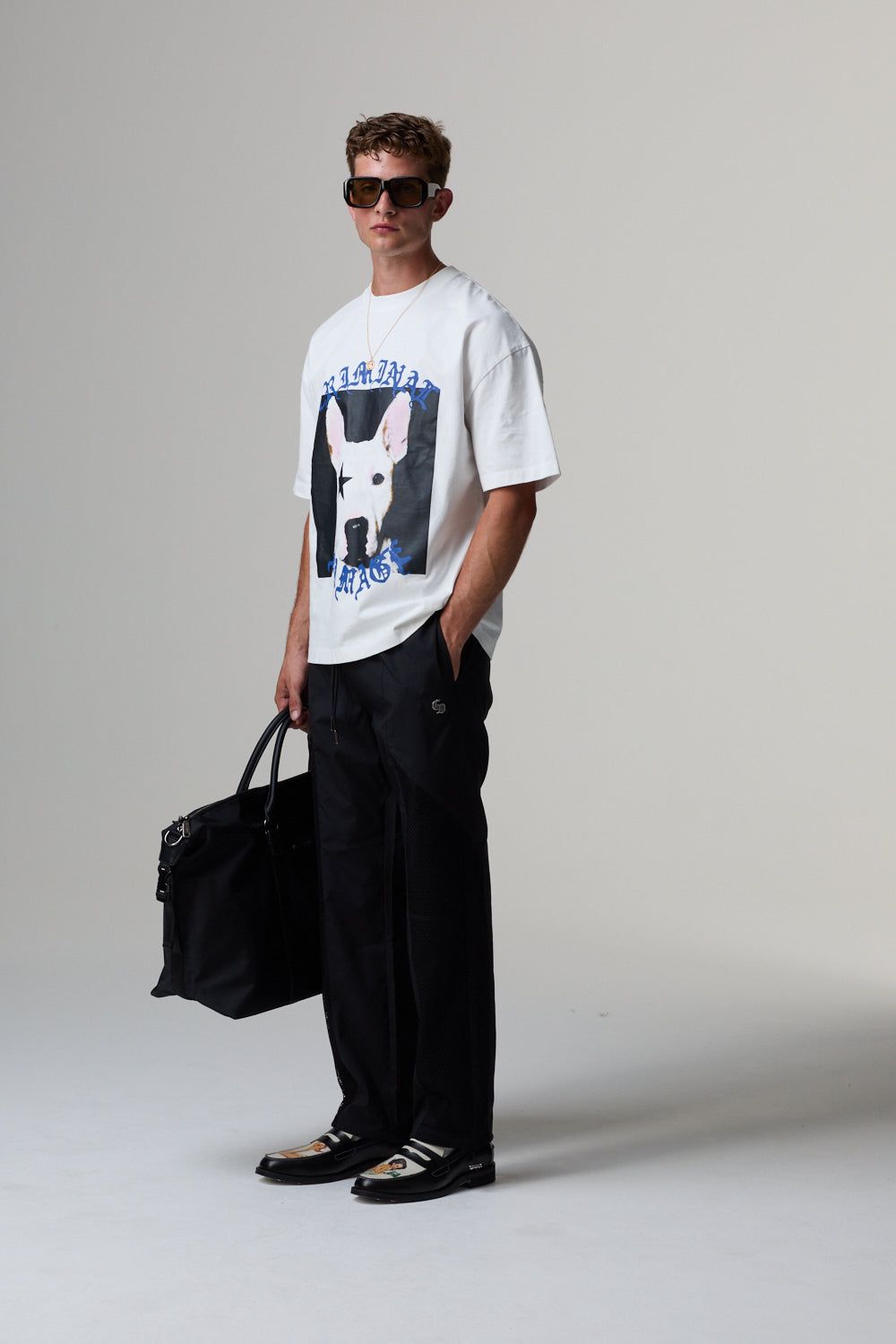 Model wearing a white Criminal Damage t-shirt with a graphic design, black pants, and sunglasses, holding a black bag on a grey background.