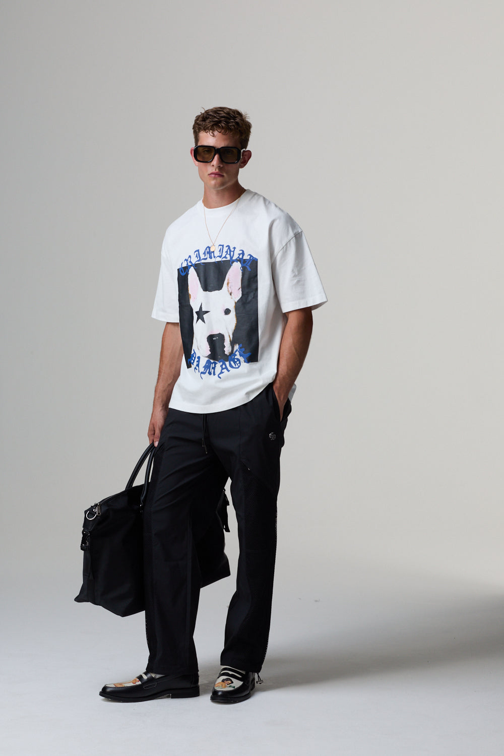 Man wearing a white Criminal Damage t-shirt with a graphic design, holding a black bag on a grey background