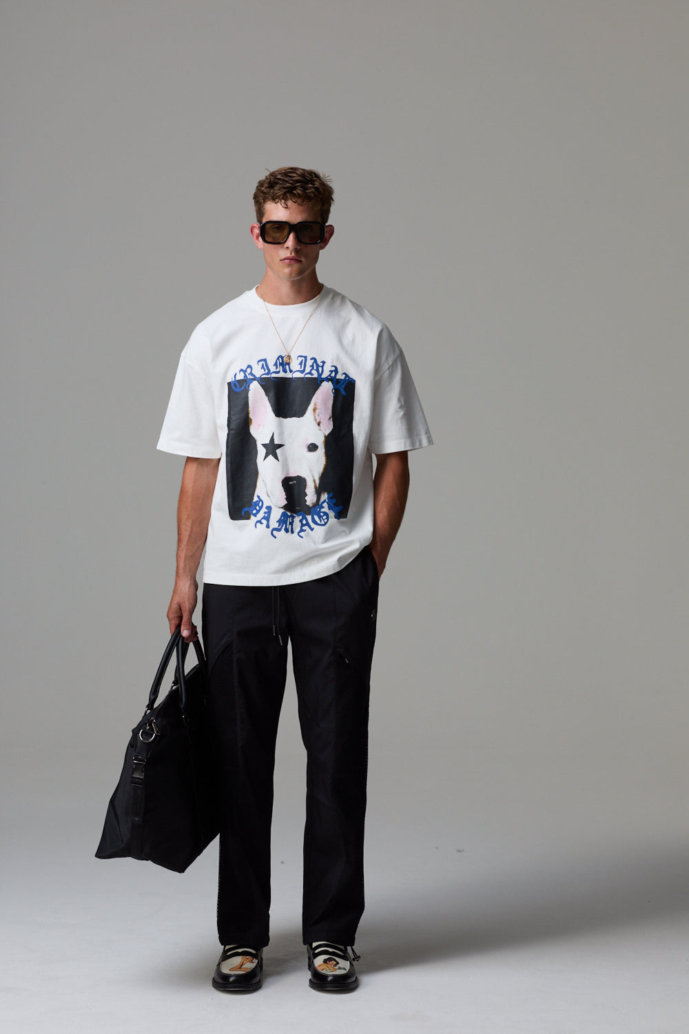 Model wearing a white Criminal Damage t-shirt with a graphic design, holding a black bag on a grey background