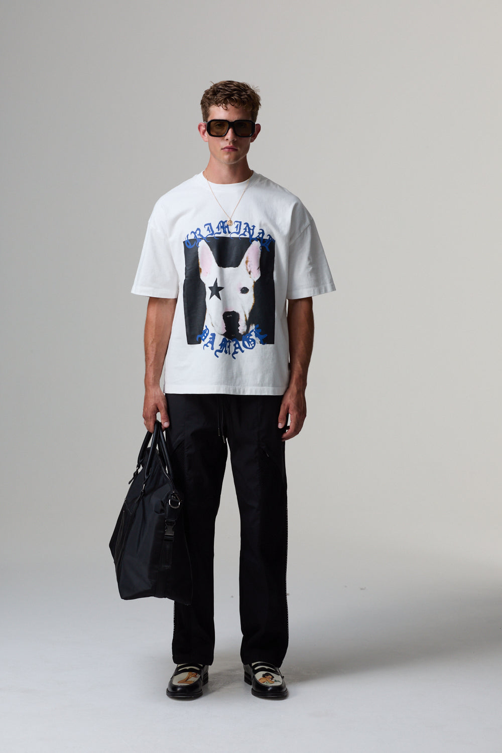 Model wearing a white Criminal Damage t-shirt with a graphic design, holding a black bag on a grey background