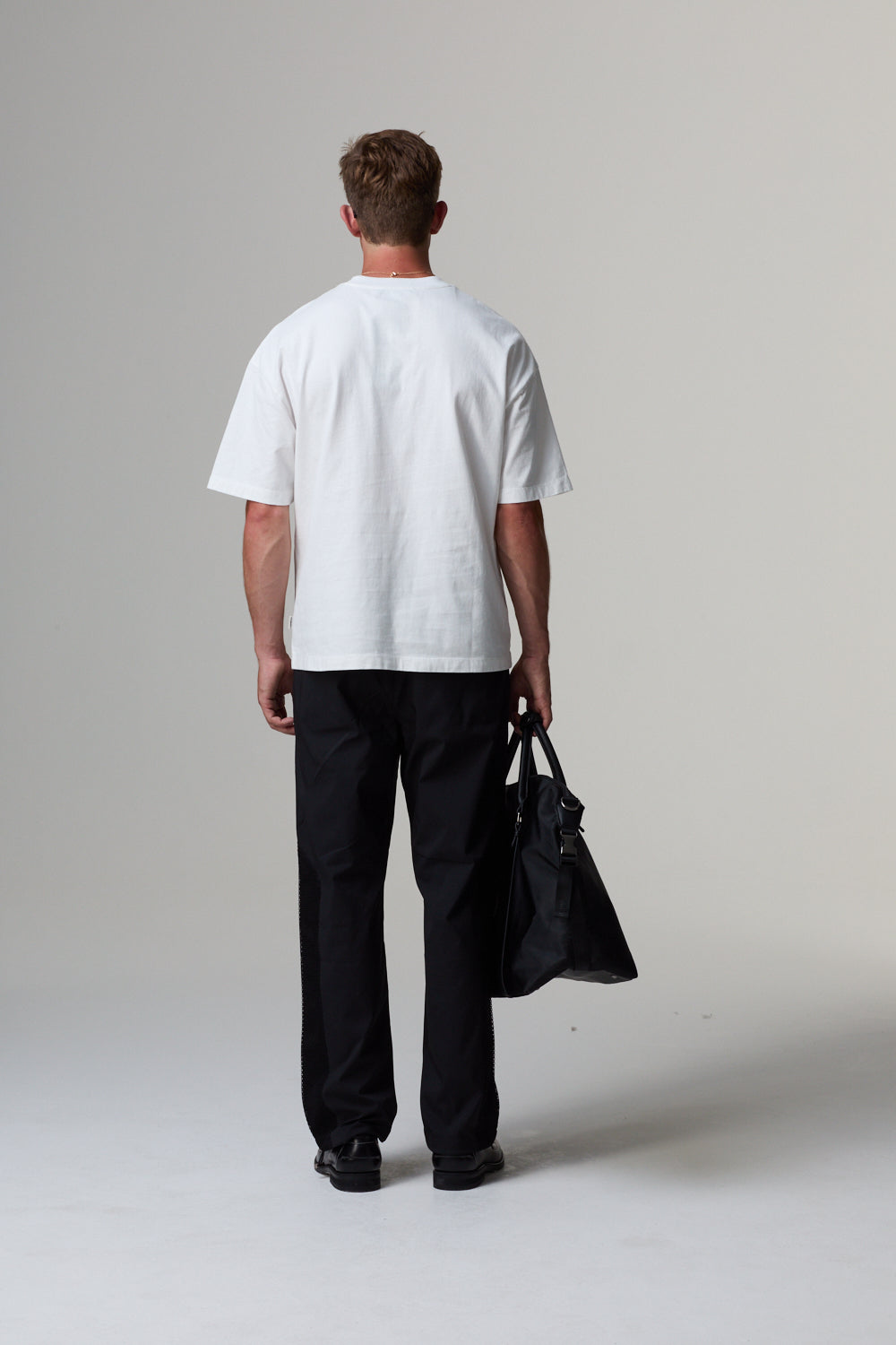 Model wearing a white Criminal Damage  t-shirt and black pants holding a black bag on a grey background