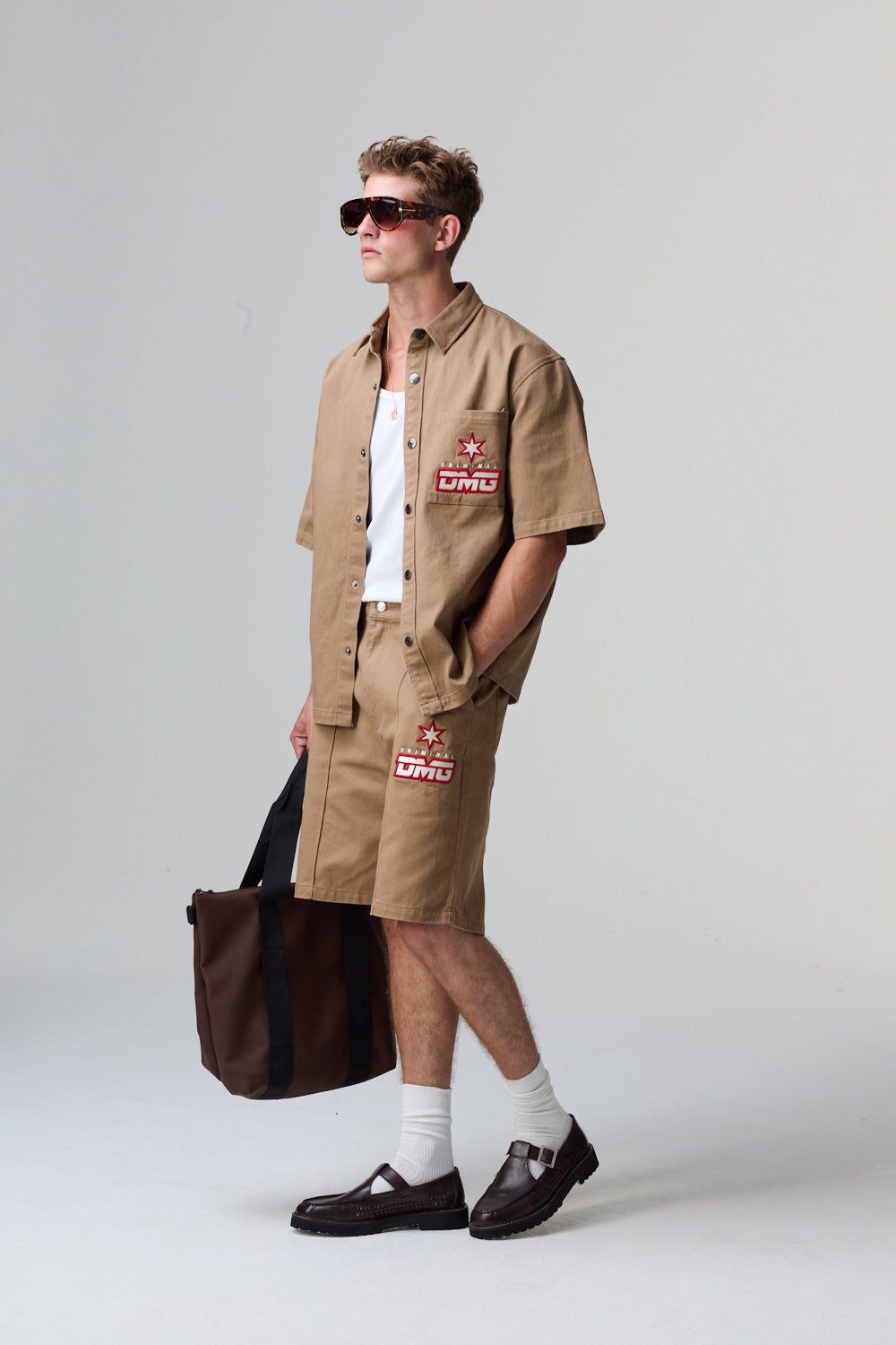 Model wearing a beige Criminal Damage shirt and shorts with patches, holding a brown bag on a grey background