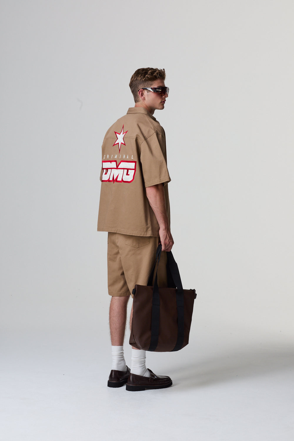 Model wearing a beige Criminal Damage outfit, holding a brown bag on a grey background