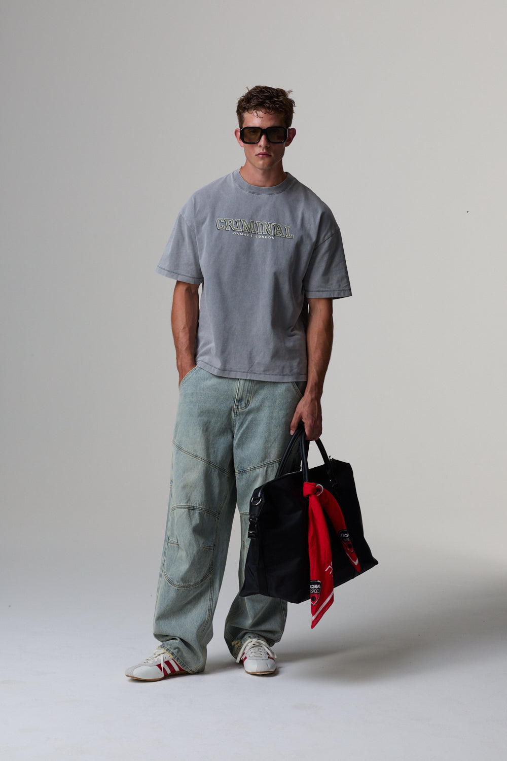 Model wearing a grey Criminal  Damage t-shirt and light blue jeans, holding a black bag.