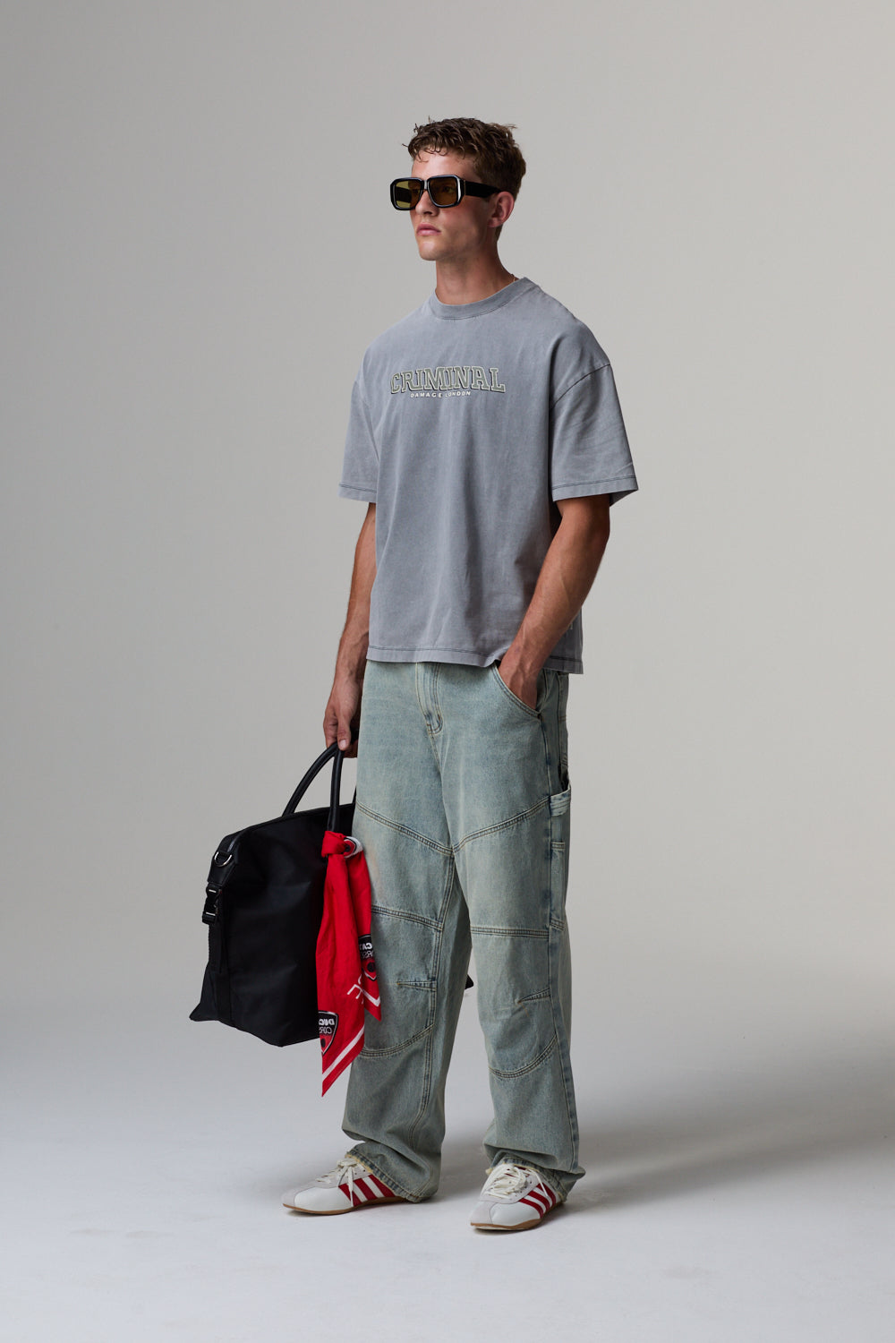 Model wearing a grey Criminal Damage t-shirt and light blue jeans, holding a black bag, on a grey background.