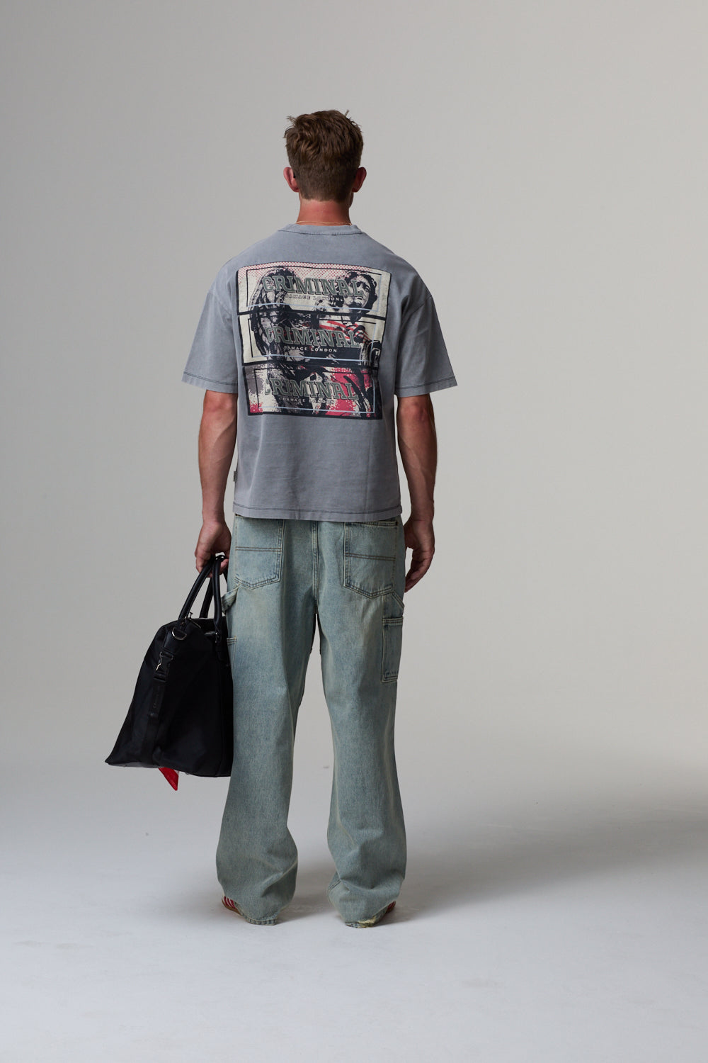 Model wearing a grey Criminal Damage t-shirt with a graphic design on the back, holding a black bag.