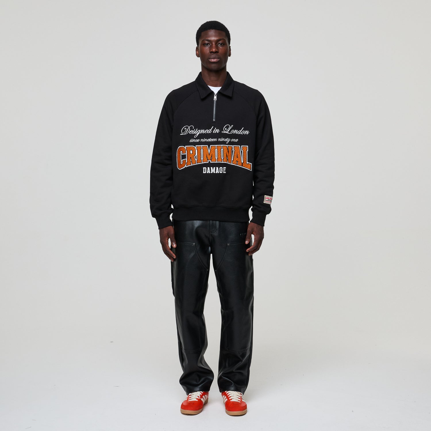 Person wearing a black Criminal Damage jacket with text, black pants, and red shoes on a white background