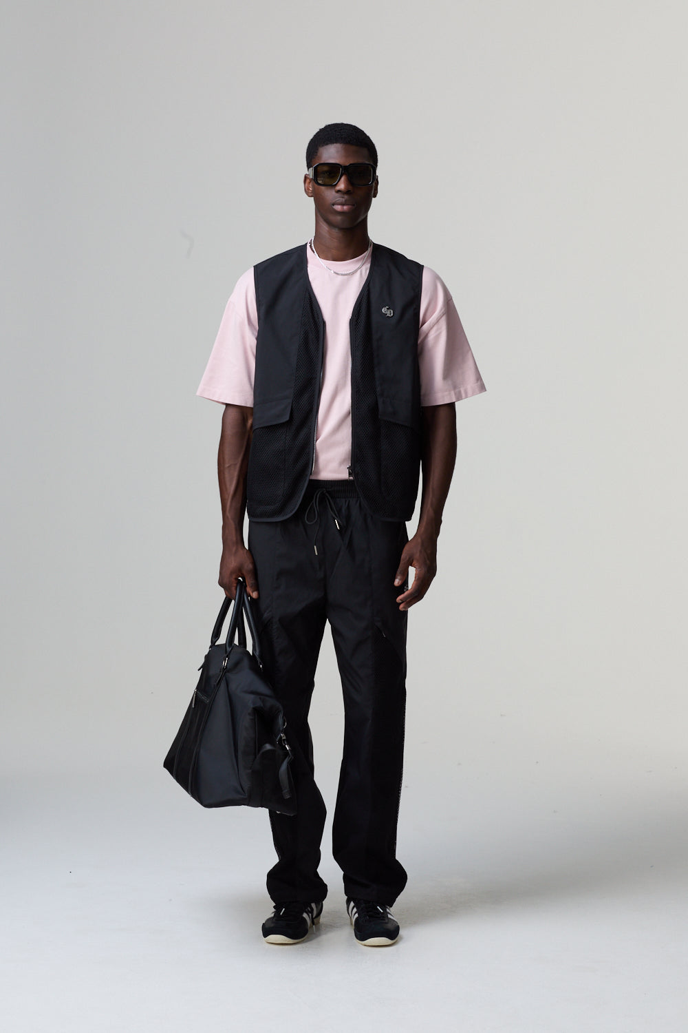 Model wearing a Criminal Damage pink shirt, black vest, and black pants holding a black bag on a grey background