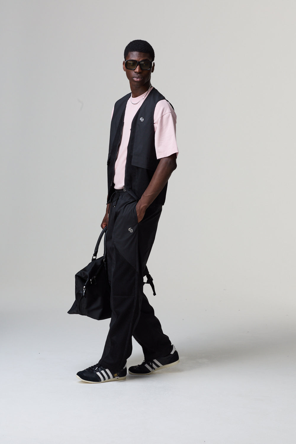 Model wearing a Criminal Damage black vest, pink shirt, and black pants holding a black bag on a grey background