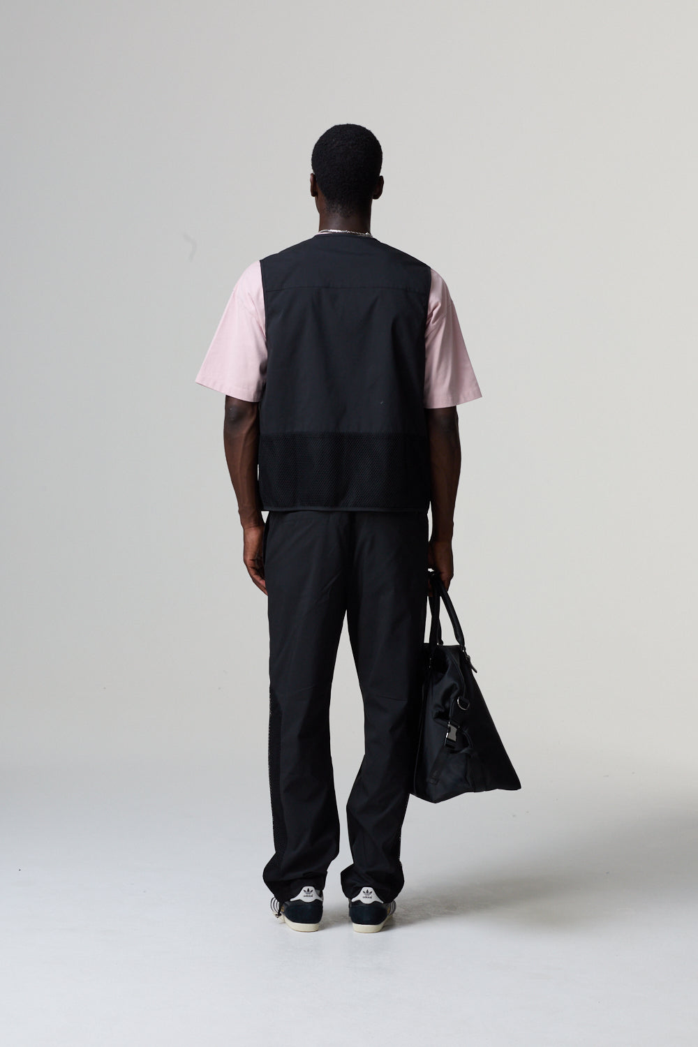 Model wearing a Criminal Damage black vest over a pink shirt with black pants, holding a black bag on a grey background