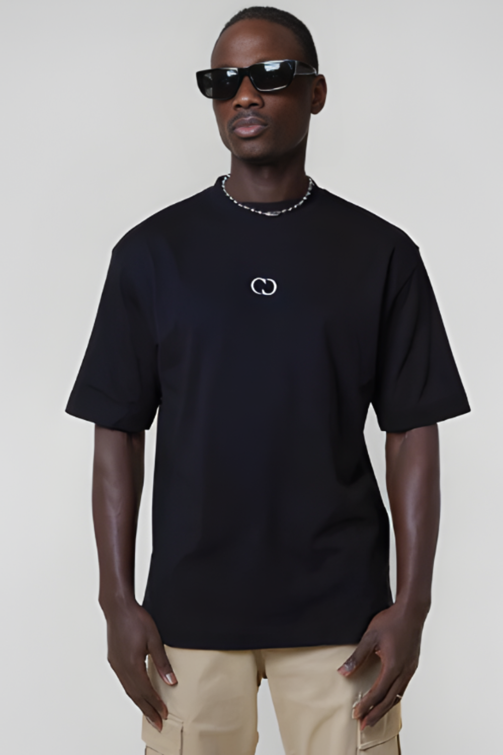 Man wearing a black t-shirt with a logo, sunglasses, and beige pants on a plain background