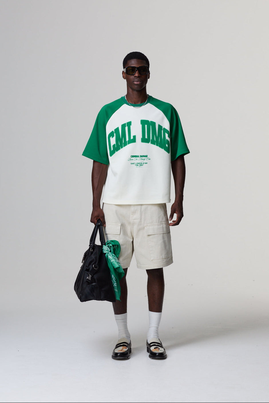 Model wearing a Criminal Damage green and white baseball shirt with 'CML LMG' text, holding a black bag on a white background
