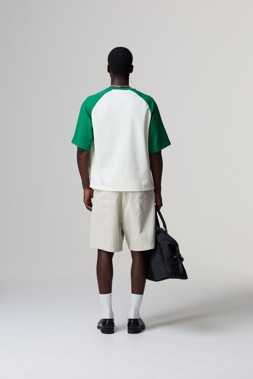 Model wearing a Criminal Damage green and white baseball shirt with beige shorts, holding a black bag on a plain background