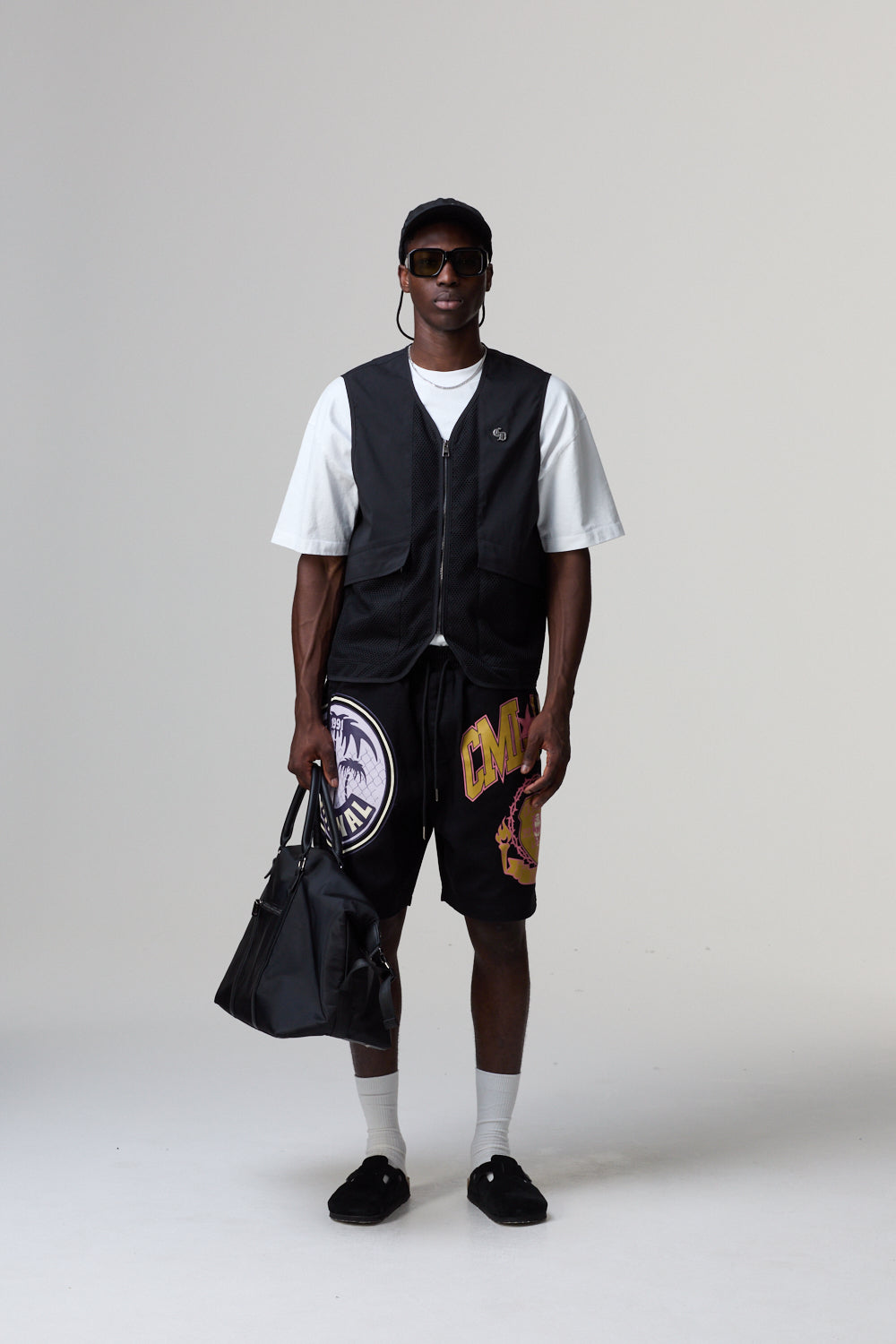 Model wearing a black Criminal Damage vest, white t-shirt, and shorts with a graphic design, holding a black bag on a grey background.
