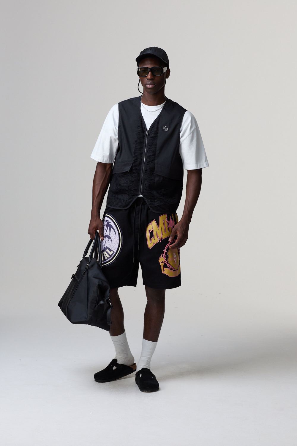 Model wearing a black Criminal Damage vest, white t-shirt, and shorts with a logo, holding a black bag on a grey background