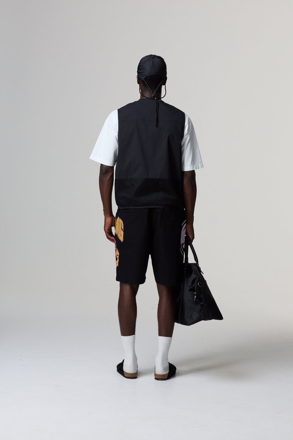 Model wearing a black Criminal Damage vest, white shirt, black shorts, and white shoes on a grey background