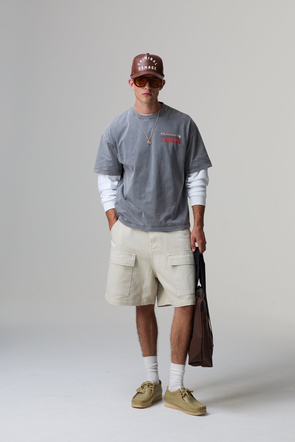 Model wearing a grey Criminal Damage sweatshirt with red text, beige shorts, and a brown cap on a grey background