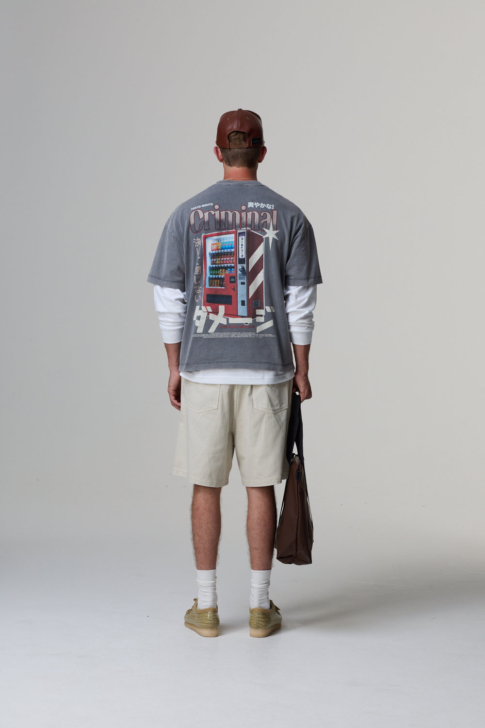 Model wearing a grey Criminal Damage t-shirt with a graphic design, beige shorts, and a brown cap on a grey background