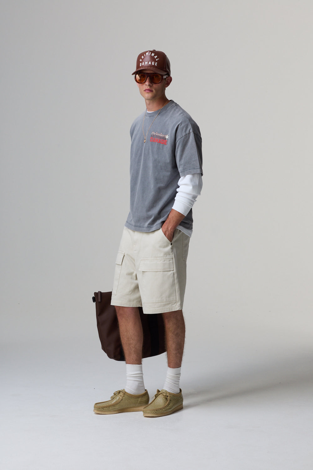 Model wearing a grey Criminal Damage t-shirt, beige shorts, and brown shoes on a grey background