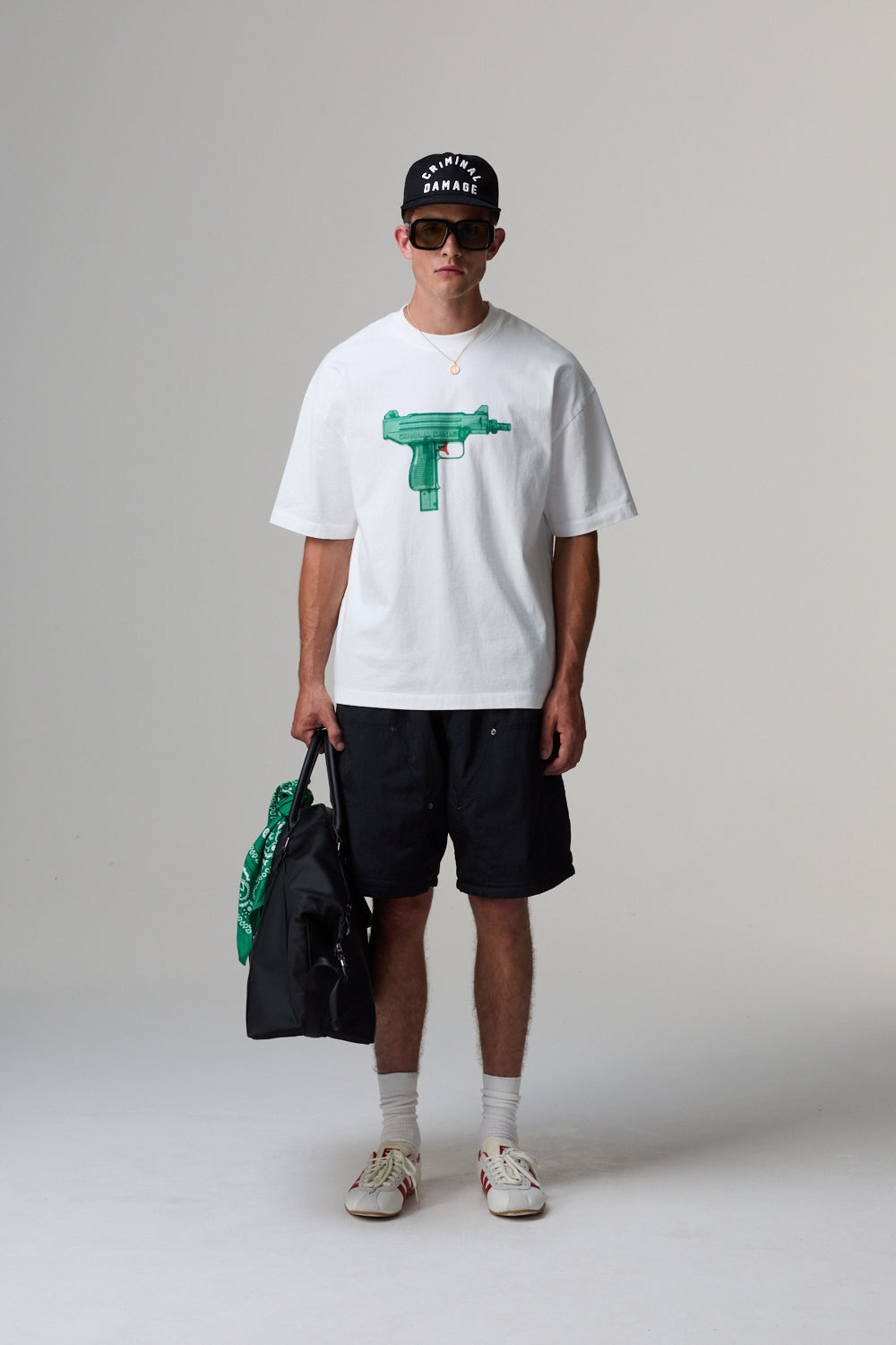 Model wearing a white Criminal Damage t-shirt with a graphic design, black shorts, and sunglasses, holding a black bag.